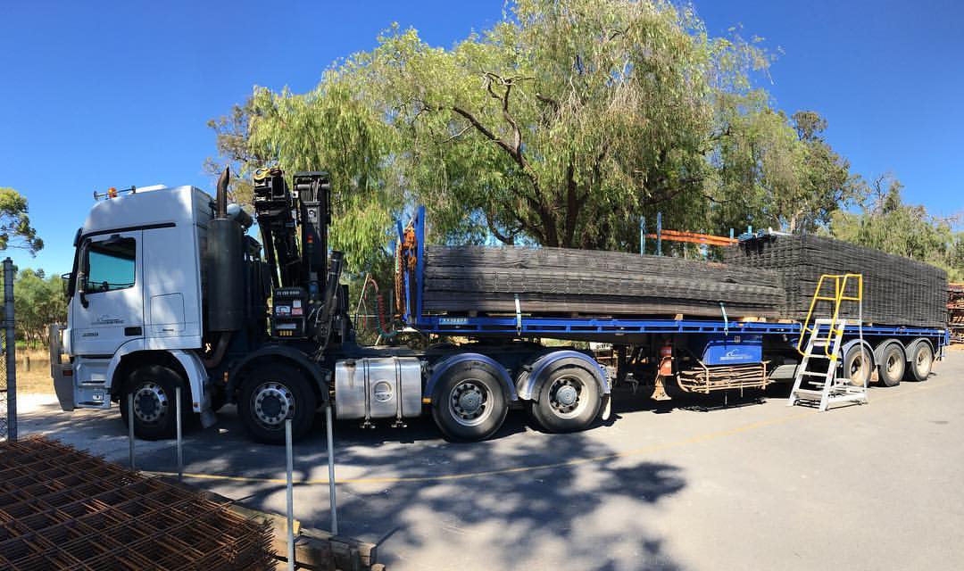 Flatbed truck carrying strapped stacks of steel reinforcing mesh beside a warehouse yard