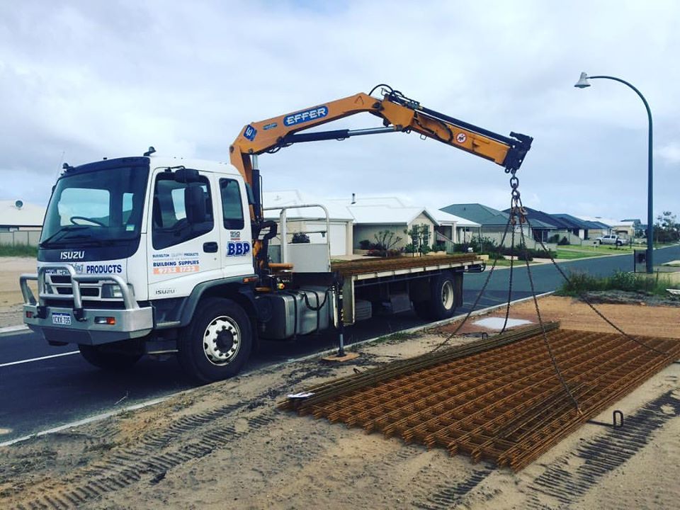 Flatbed truck with an extended hydraulic crane lifting steel reinforcing mesh beside a residential street